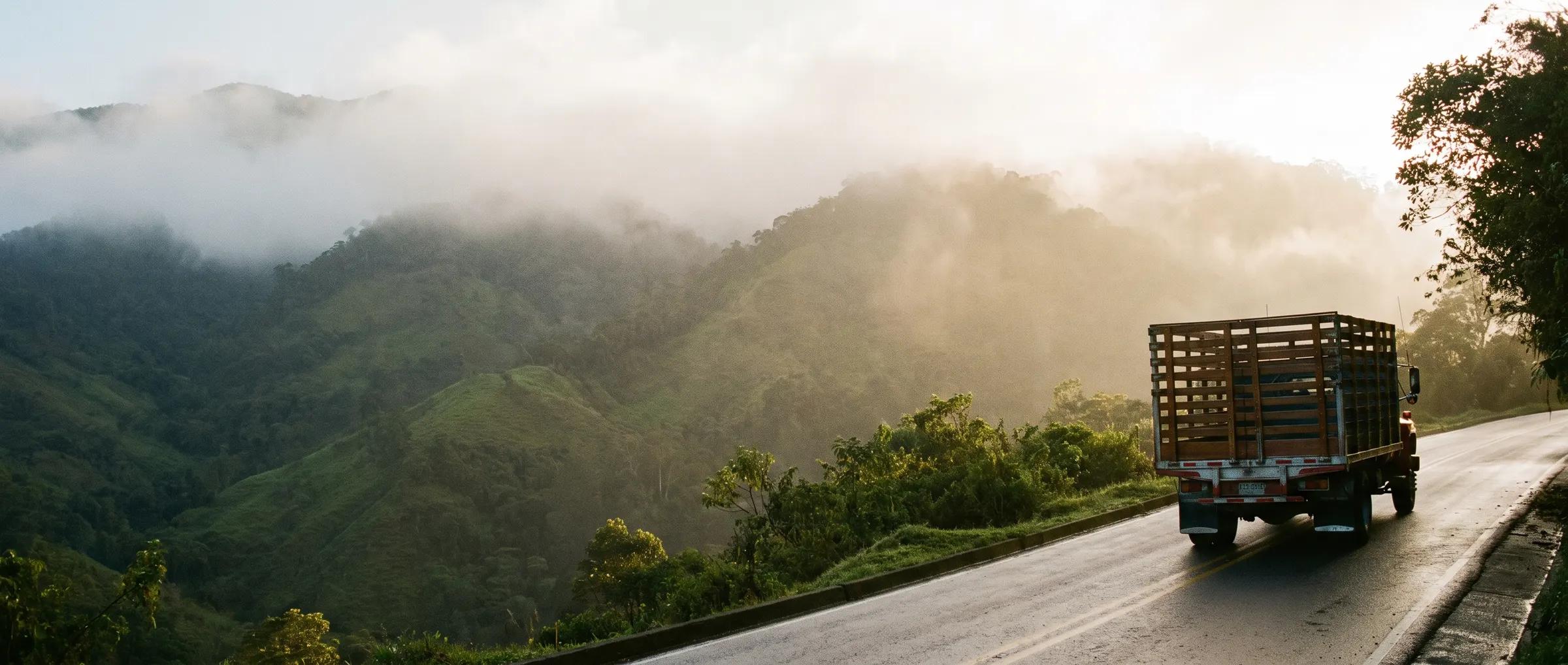 Un camión de estacas sube una carretera de montaña colombiana al amanecer, con niebla sobre la cordillera.